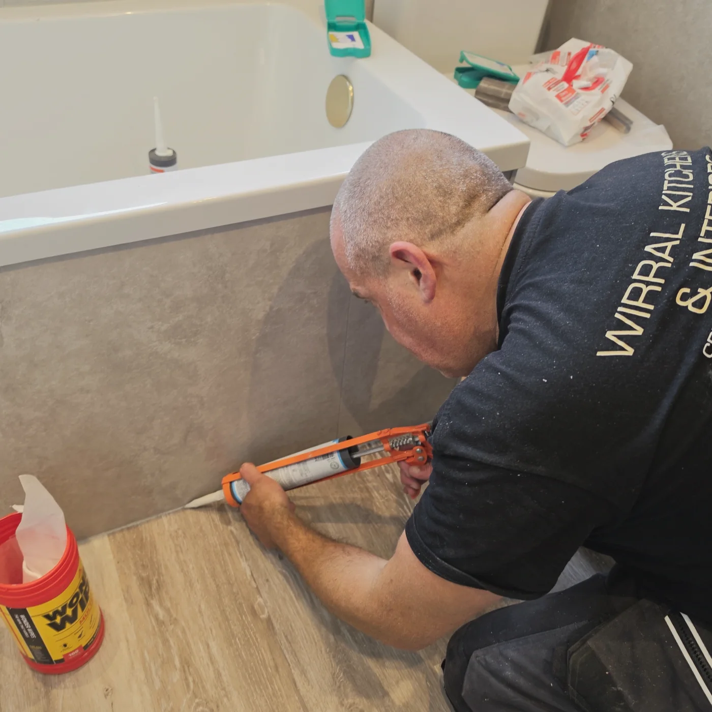 A tradesperson in Wirral Kitchens & Interiors workwear applying fresh sealant along the edge of a bath.